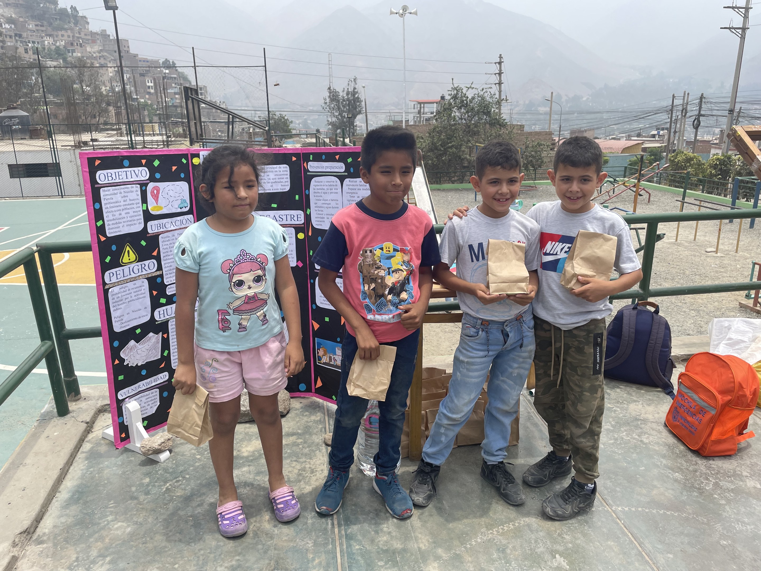 Gruppe von Kindern auf einem Basketballfeld mit Papiertüten in den Händen, Tüten und Gegenstände rechts daneben und eine Tafel mit Text links daneben, vor einem Hintergrund aus Zäunen, Pfosten, Straßenlaternen, Bäumen, Gebäuden, Hügeln und Himmel.