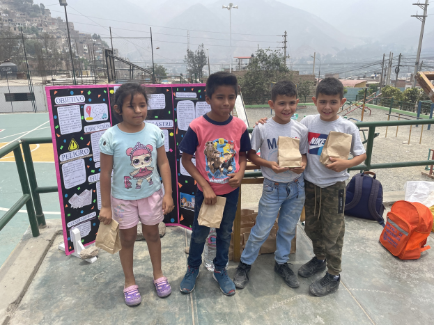 Gruppe von Kindern auf einem Basketballfeld mit Papiertüten in den Händen, Tüten und Gegenstände rechts daneben und eine Tafel mit Text links daneben, vor einem Hintergrund aus Zäunen, Pfosten, Straßenlaternen, Bäumen, Gebäuden, Hügeln und Himmel.