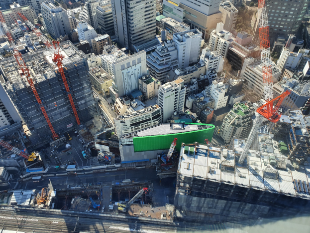 Luftaufnahme einer Stadt mit hohen Gebäuden, Kränen, Straßen, Fahrzeugen und Bäumen, die eine große grüne Baustelle in der Mitte zeigt.