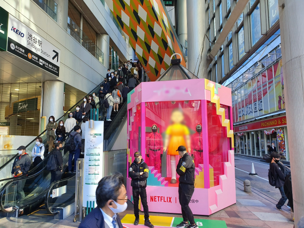 Eine Gruppe von Menschen steht vor einem modernen Gebäude mit Glaswänden, Säulen und Texttafeln; einige tragen Masken, und eine Rolltreppe mit Menschen ist zu sehen. In der Mitte befindet sich eine pinkfarbene Box mit Schaufensterpuppen und dem Text "Netflix's new Netflix booth in Tokyo."