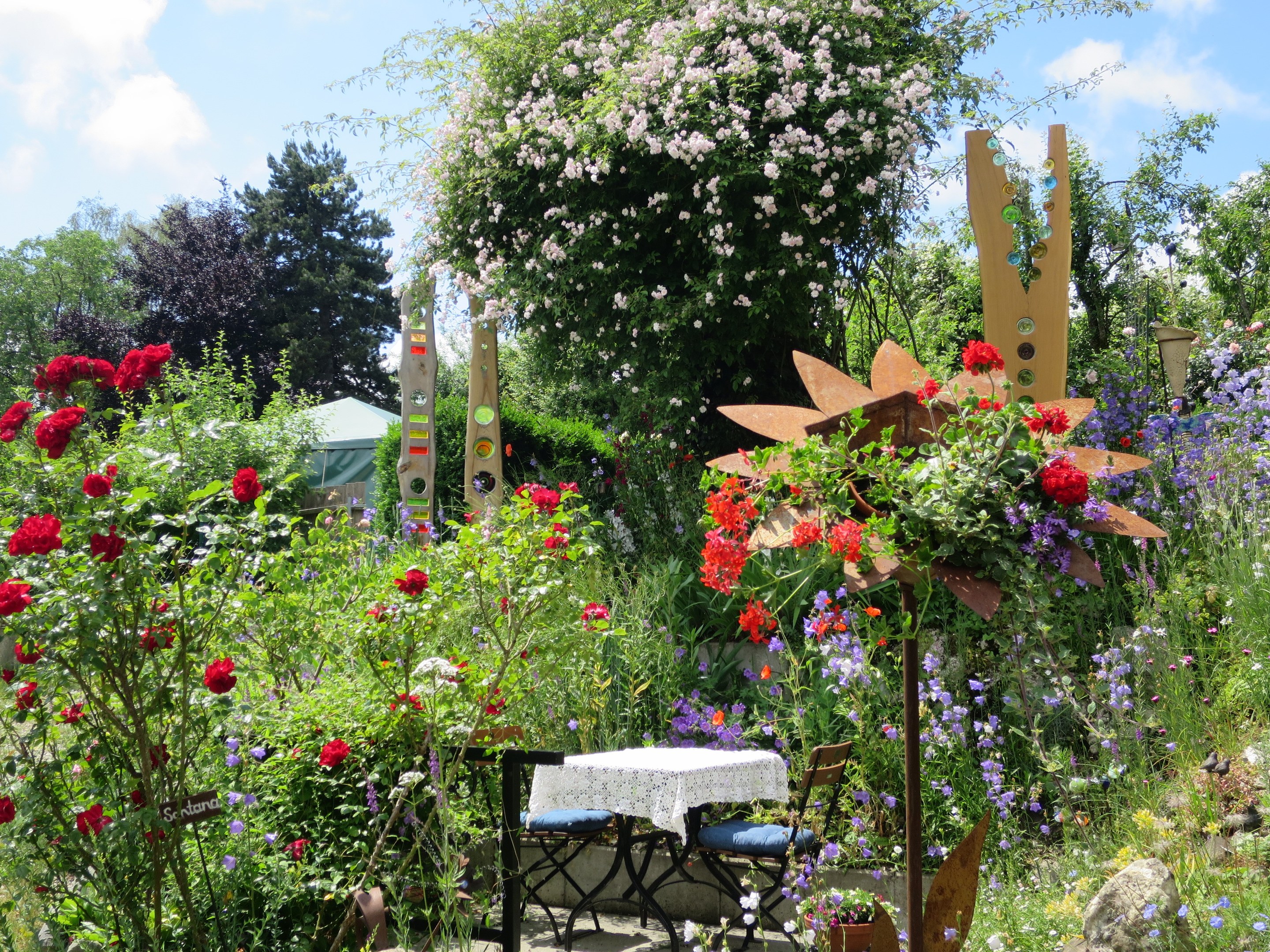 Ein Garten mit bunten Blumen und Pflanzen, der einen Tisch mit einer Decke und Stühlen in der Mitte zeigt, umgeben von Bäumen unter einem bewölkten Himmel auf der Chelsea Flower Show.