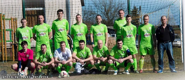 Eine Gruppe von Männern in grünen T-Shirts steht auf einem Fußballfeld mit einem Ball davor, einige in der Hocke, in der Nähe eines Torpfostens, mit Bäumen, Gebäuden und Fahrzeugen im Hintergrund.