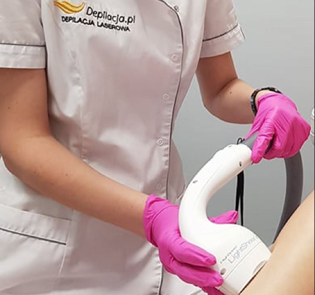 Eine Frau in einem weißen Hemd und pinken Handschuhen bedient eine weiße Laser-Haarentfernungsmaschine an ihrer Haut, vor einer Wand stehend.