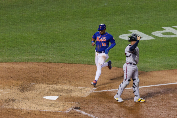 Baseballspieler in Uniform und Helm läuft Richtung Homeplate, hält einen Schläger, auf einem grasbedeckten Feld mit Text am Boden.