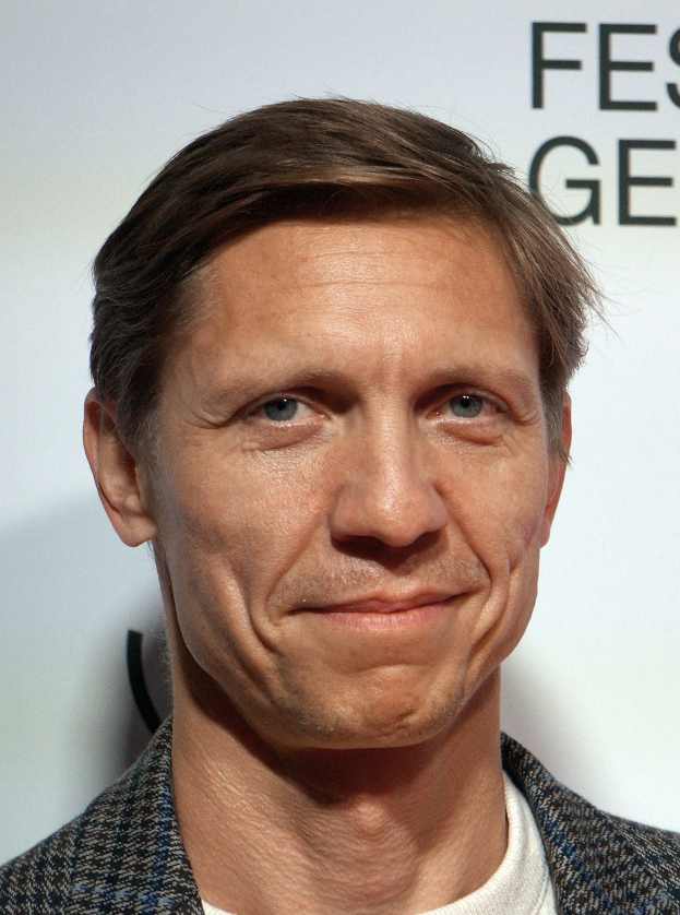 Ein Mann im Anzug lächelt in den Vordergrund, während im Hintergrund eine Tafel mit 'Eddie Redmayne Net Worth' zu sehen ist.