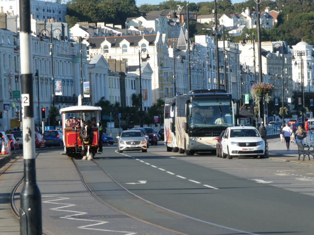 Eine belebte Stadtstraße mit hohen Gebäuden, Fahrzeugen, Fußgängern, Verkehrsampeln, Laternenmasten, Schildern, Bäumen und einem klaren blauen Himmel.