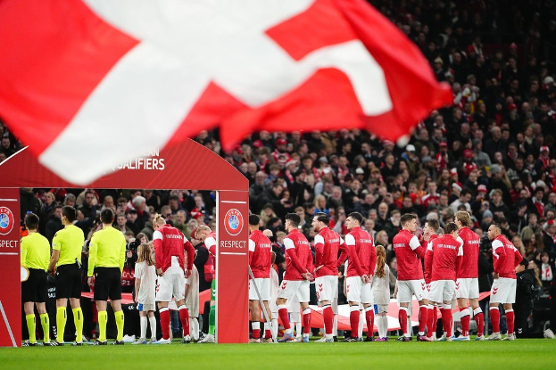 Gruppe von Menschen auf einem Fußballfeld mit einer roten und weißen Fahne im Vordergrund, einem Bogen mit der Aufschrift "Bayern München vs Bayern München Wett-Tipps & Vorschau" im Hintergrund und einer großen Stadionmenge.