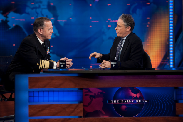 Zwei Männer in schwarzen Blazern, Hemden und Krawatten sitzen an einem Tisch mit Mikrofonen zwischen ihnen und unterhalten sich vor einem Fernseher und einem blauen Hintergrund.