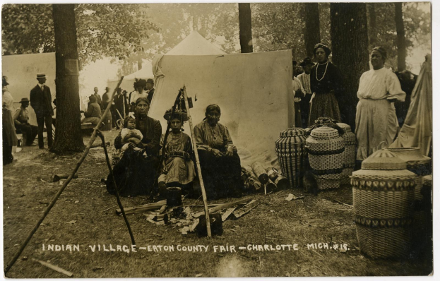 Schwarz-weiß-Foto einer Gruppe von Menschen, die um ein Zelt auf dem Eton County Fair in Charlotte, North Carolina sitzen und stehen, mit Körben und Gegenständen auf der rechten Seite, Bäumen und einer Wand im Hintergrund und Text unten.