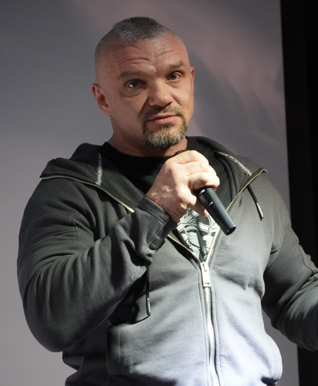 Ein Mann in einer schwarzen Kapuzenjacke und einem schwarzen Anzug mit einem weißen Hemd, zurückgekämmten Haaren und einem selbstbewussten Gesichtsausdruck hält ein Mikrofon, während er vor einer Wand steht.