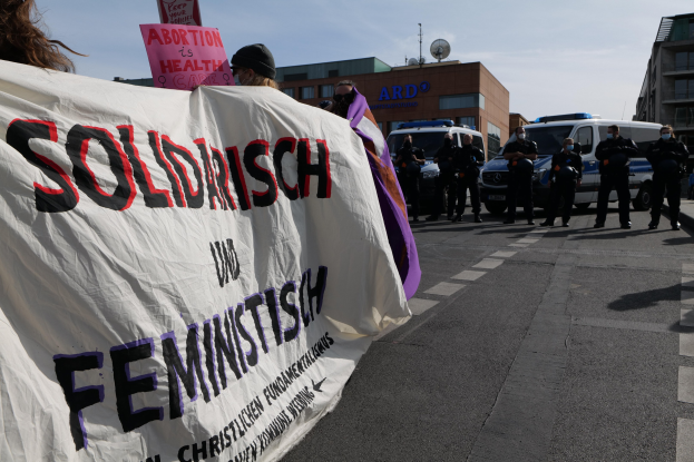 Eine Gruppe von Menschen marschiert auf einer Straße, trägt ein Banner mit der Aufschrift 'Solidarität und Feminismus', mit parkenden Fahrzeugen und Gebäuden im Hintergrund unter einem klaren blauen Himmel.