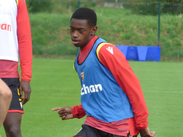 Ein junger Mann in einem blauen T-Shirt und schwarzen Shorts, der auf einem grünen Feld mit einem Zaun, Pflanzen und Bäumen im Hintergrund Füßball spielt.