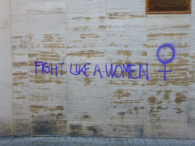 Eine Wand mit den Worten "Kämpfe wie eine Frau" in blauem Graffiti, mit einem Fenster oben und einem Boden unten.