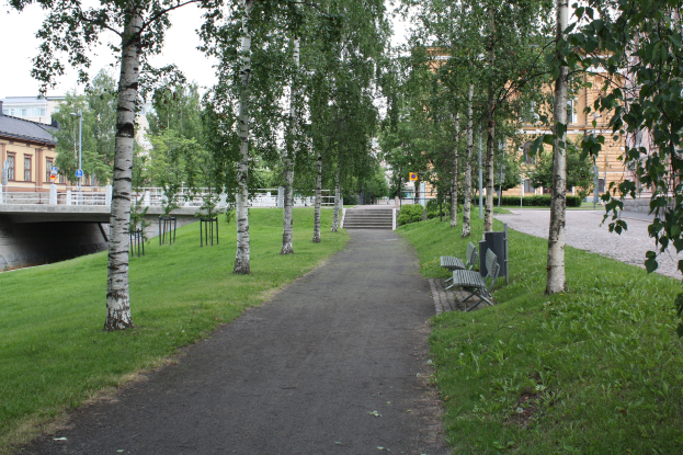 Ein Park mit einem baumbestandenen Weg, Bänken, Gras, einer Brücke mit Geländer, Gebäuden im Hintergrund, Treppen, Laternenpfählen und Himmel.