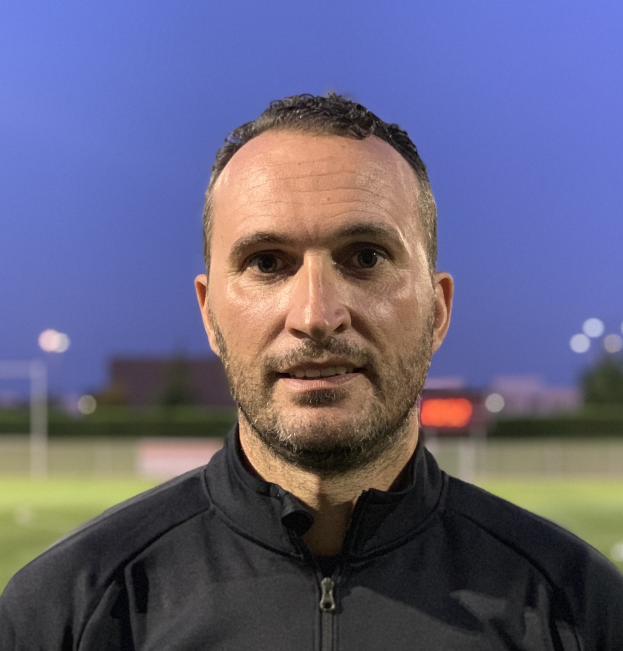 Ein Mann in einer schwarzen Jacke mit einem Logo steht auf einem Fussballfeld und scheint ein Trainer zu sein.