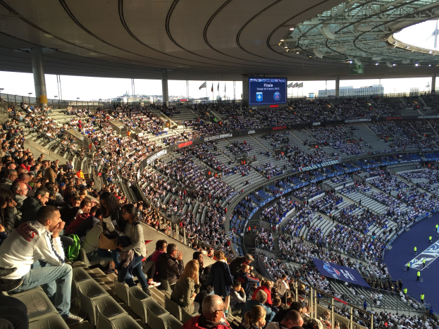 Große Menschenmenge im Allianz Arena Stadion bei einem Fußballspiel sitzend, mit einer Bühne rechts, Fahnen, Stangen und einem Bildschirm im Hintergrund.