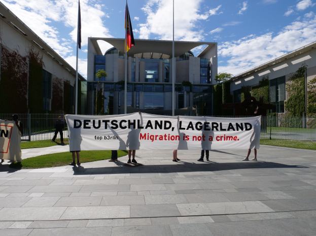 Gruppe von Menschen mit einem Transparent mit der Aufschrift 'Deutschland, Lagerland Migration ist kein Verbrechen' vor einem Gebäude mit Fenstern, umgeben von Gras, einem Metallzaun, Pflanzen, Bäumen und Fahnen mit Stangen unter einem bewölkten Himmel.