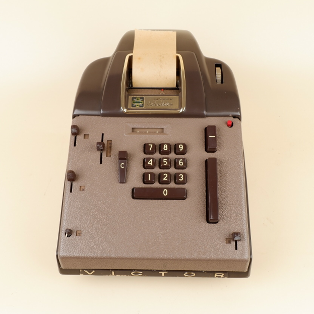 Eine altmodische Kasse mit schwarzer und silberner Farbgebung, die einen großen Bildschirm, eine Tastatur und mehrere Knöpfe und Drehregler aufweist, steht auf einem weißen Tisch.