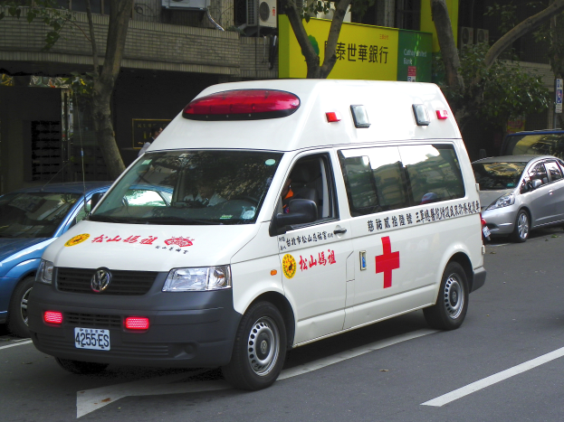Ein Krankenwagen steht auf der Seite einer Straße, umgeben von anderen Fahrzeugen, mit Bäumen, Gebäuden, Texttafeln und Klimaanlagen im Hintergrund.