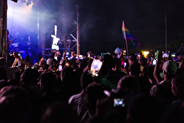 Eine Menge bei einem Konzert mit hochgehaltenen Handys, beleuchtet von Lichtern, umgeben von Pfosten und einer Flagge, während Musiker auf der Bühne spielen.