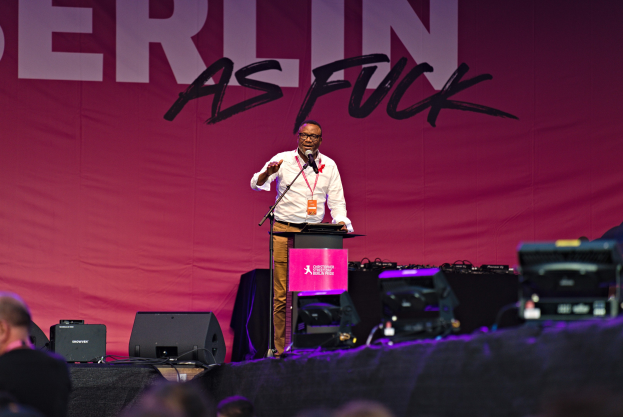 Mann in weißem Hemd und braunen Hosen, der in ein Mikrofon spricht, vor einem Publikum auf einer Bühne mit einem pinken 'Berlin as Fuck'-Plakat dahinter.