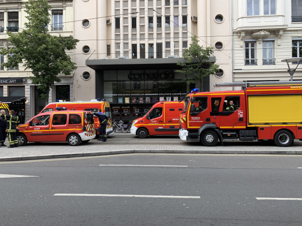 Eine Gruppe von Feuerwehrautos auf einer Pariser Straße geparkt, mit Menschen in der Nähe, vor Häusern, Bäumen und einem Fahrrad, wahrscheinlich am Unfallort.