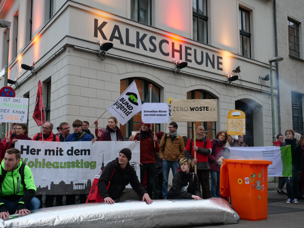 Menschen mit Schildern und Plakaten stehen vor einem Gebäude während einer Demonstration in Deutschland, wobei zwei Personen im Vordergrund sitzen und ein Müllcontainer rechts zu sehen ist.