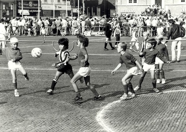 Schwarzes Foto von jungen Jungs, die auf der Straße Fußball spielen, umgeben von einer Zuschauermenge hinter einem Geländer, mit Gebäuden, Pfählen und Texttafeln im Hintergrund.