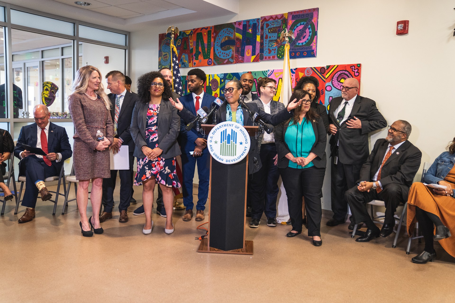 Gruppe von Menschen vor einem Podium mit Mikrofon und einer Tafel mit der Aufschrift "Sancho City Councilman Joins City Council", mit sitzenden und stehenden Teilnehmern, Flaggen, einem Feuerlöscher und Deckenleuchten im Hintergrund.