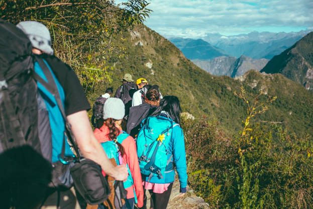 Gruppe von Menschen wandert einen Berg hinauf, mit Rucksäcken und Hüten, unter einem bewölkten Himmel mit Bäumen und Hügeln in der Landschaft.
