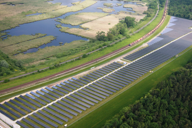 Luftbild einer Solar-Farm mit Solar-Panelen, umgeben von Bäumen, Gras, Pflanzen und Wasser, mit einem Zug auf einer nahen Bahnstrecke.